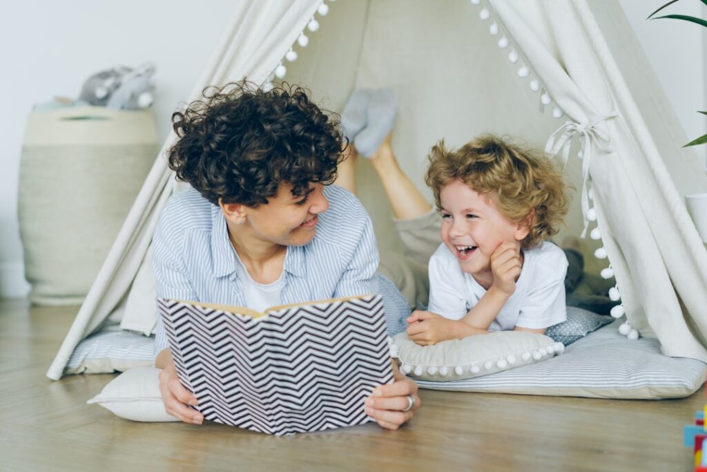 A caregiver and young child sit together reading a colorful picture book in a bright, cozy living room.