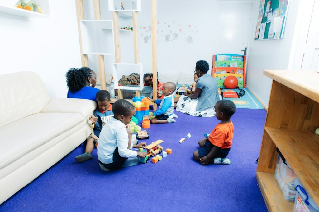 Preschool children build with blocks while a teacher kneels nearby in a bright classroom.