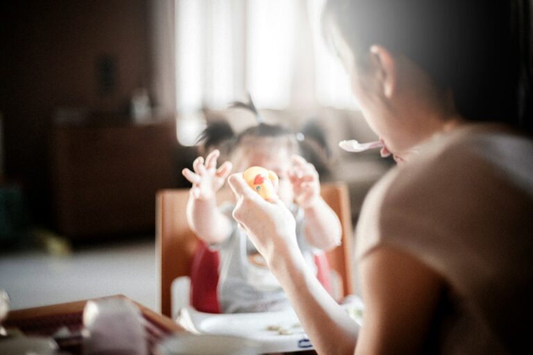 A young child explores sliced fruit and vegetables at a family table while an adult offers gentle encouragement.