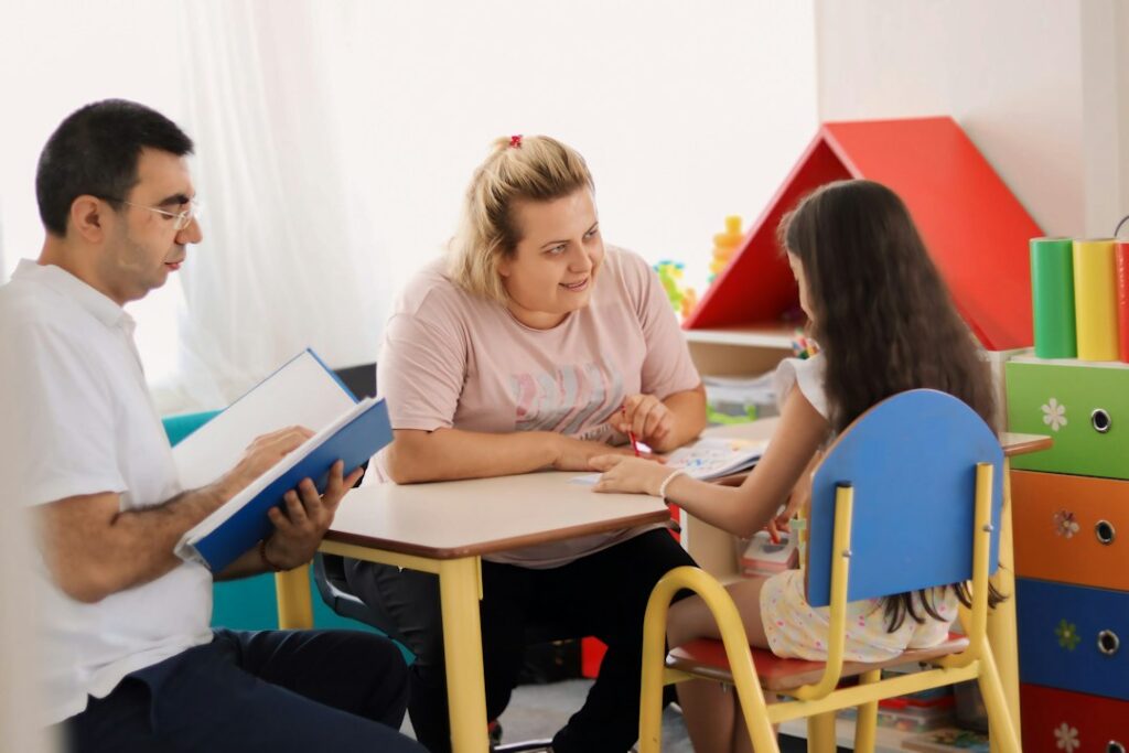 A preschool teacher and parent discuss a child’s artwork and classroom progress at a small table.