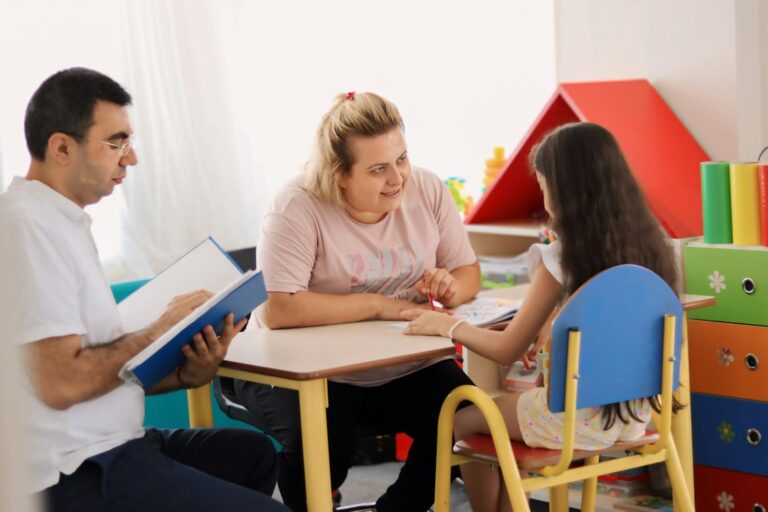 A preschool teacher and parent discuss a child’s artwork and classroom progress at a small table.